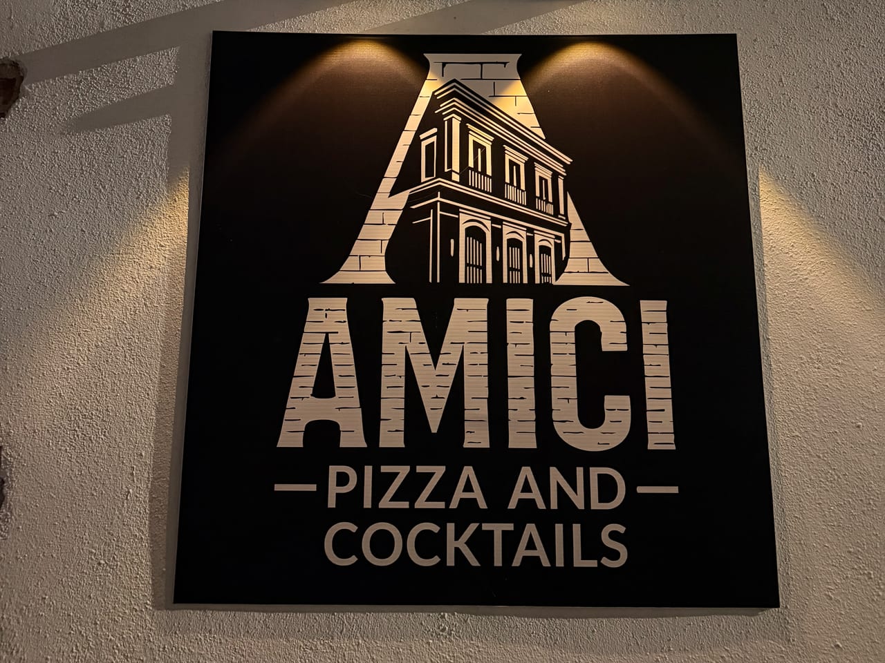 The lit wall sign outside Amici: a big letter A framing a two-story colonial building, over the words AMICI, Pizza and Cocktails